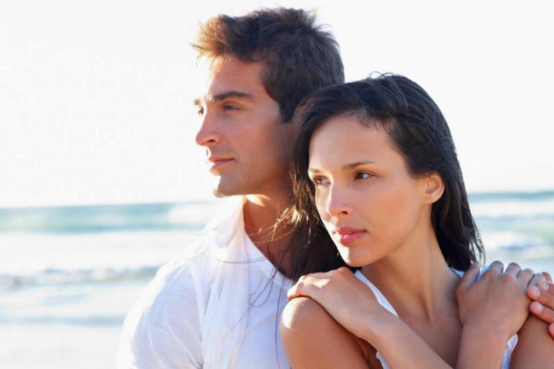 A couple standing on a beach, the woman leaning into the man's chest, her arms around herself, one of his hands on her shoulder.