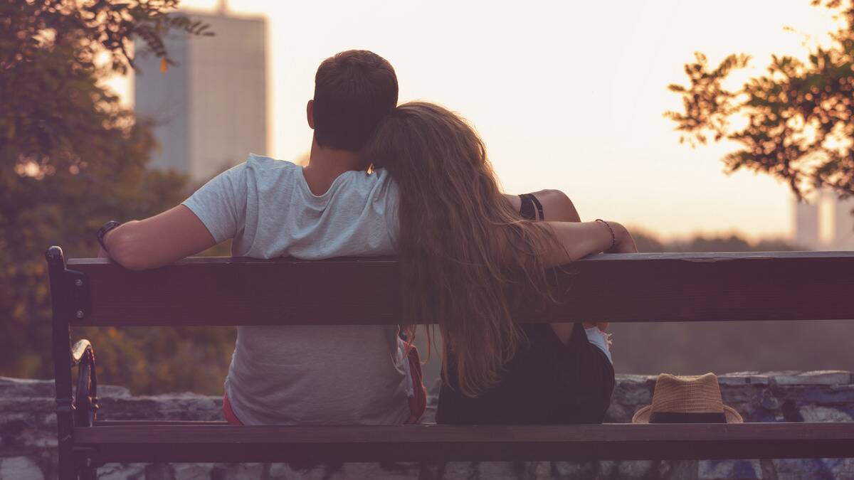 A photo from behind of a couple sitting on a bench, the woman leaning on the man's shoulder, his arm around her.
