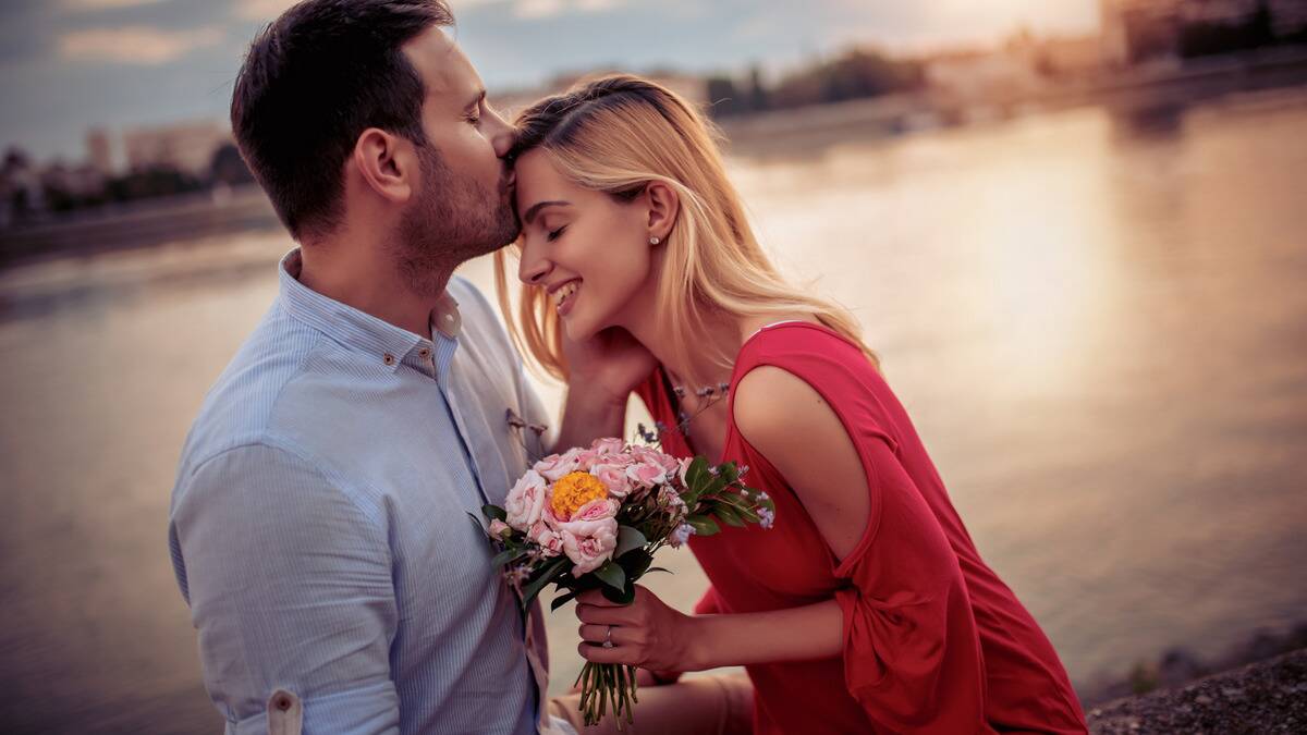 A couple standing by the water, the woman holding a bouquet of flowers, the man kissing her forehead and putting his hand on her jaw.