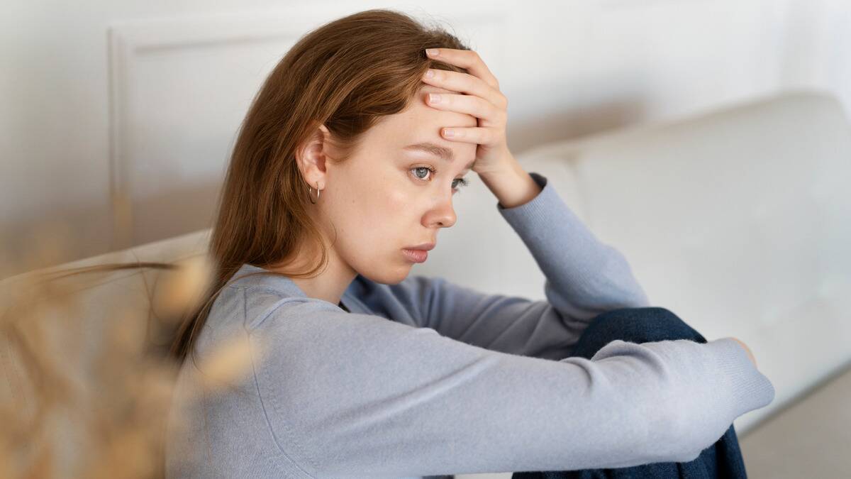 A woman sitting on a couch with a hand on her forehead, looking sad.