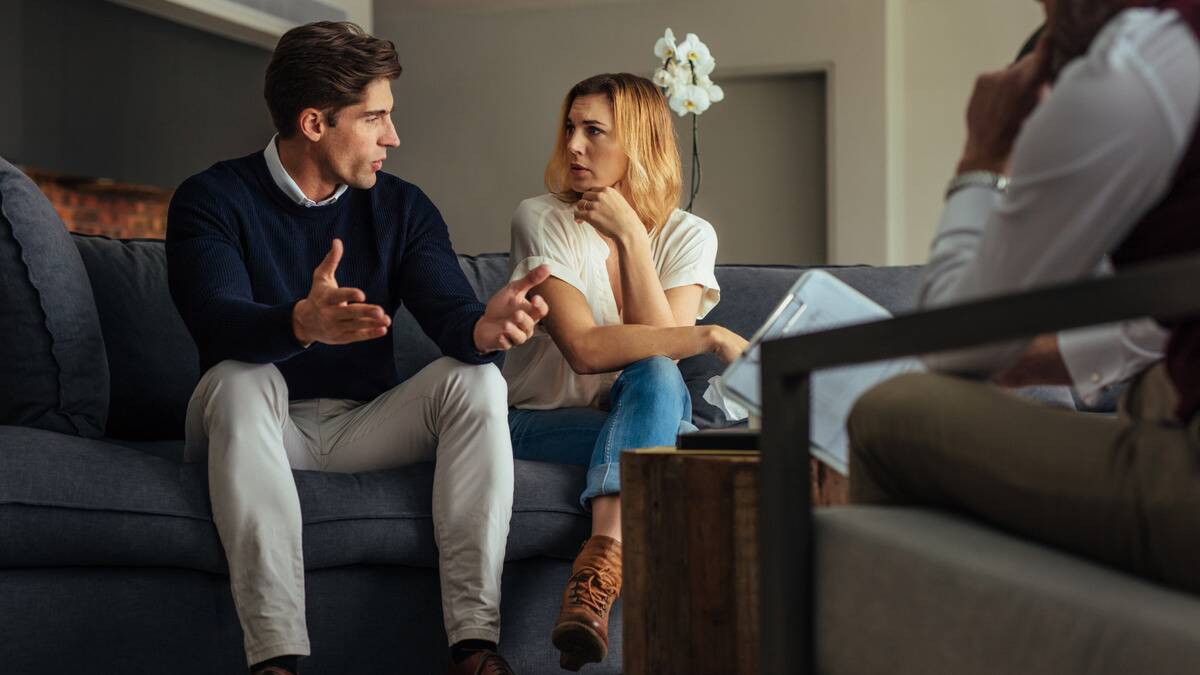 A couple sitting next to one another on a couch, talking as a couple's therapist sits across from them.