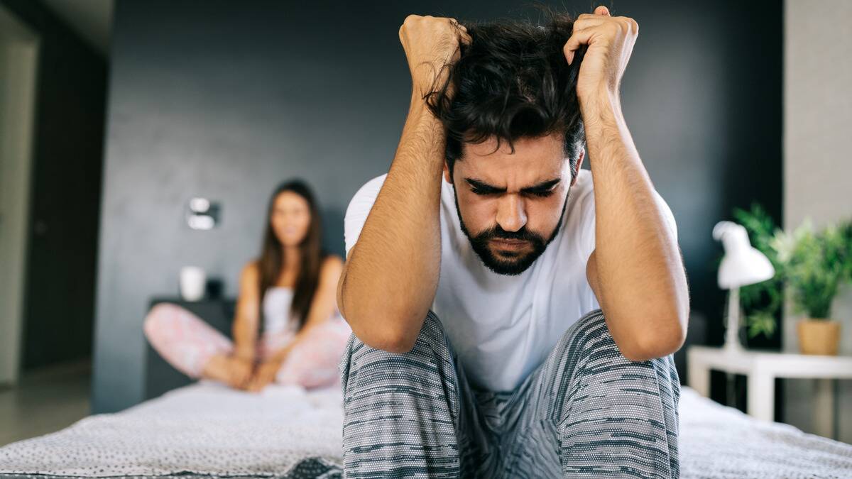 A man sitting on the edge of his bed, frustrated, hands in his hair and knees to his chest, as his girlfriend sits by the headboard behind him.