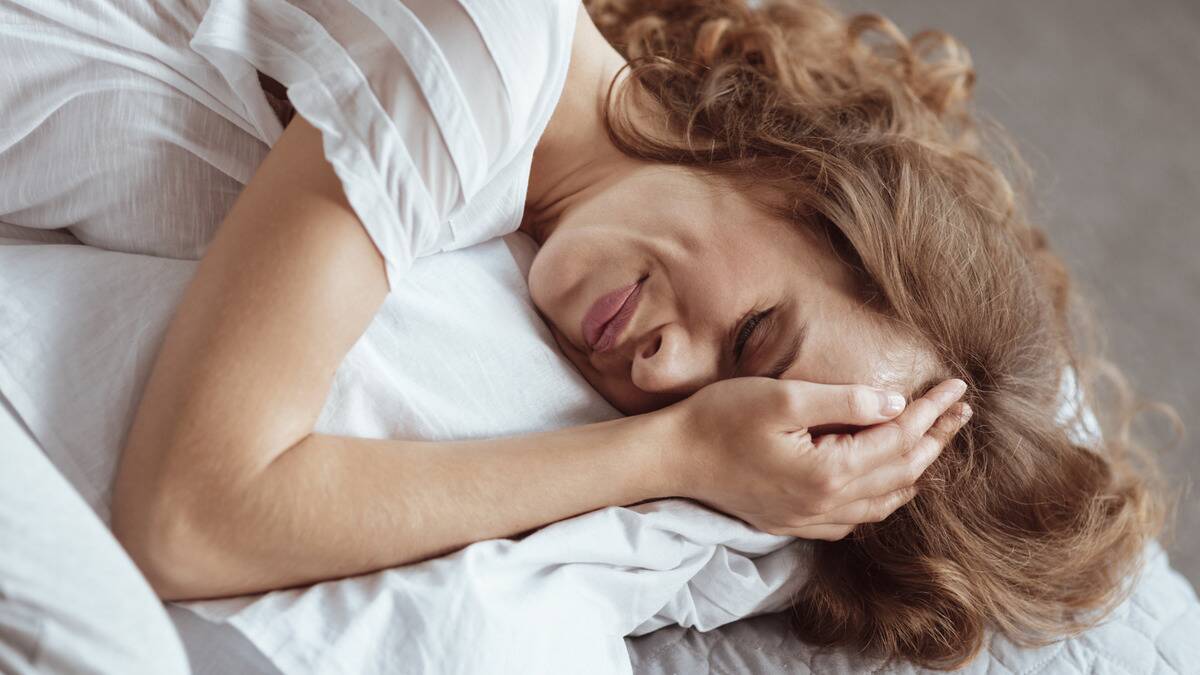 A woman laying in bed, hand on her forehead, looking pained.