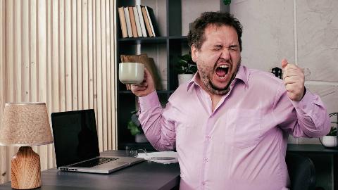 A man yelling at his desk, holding a mug in one hand.