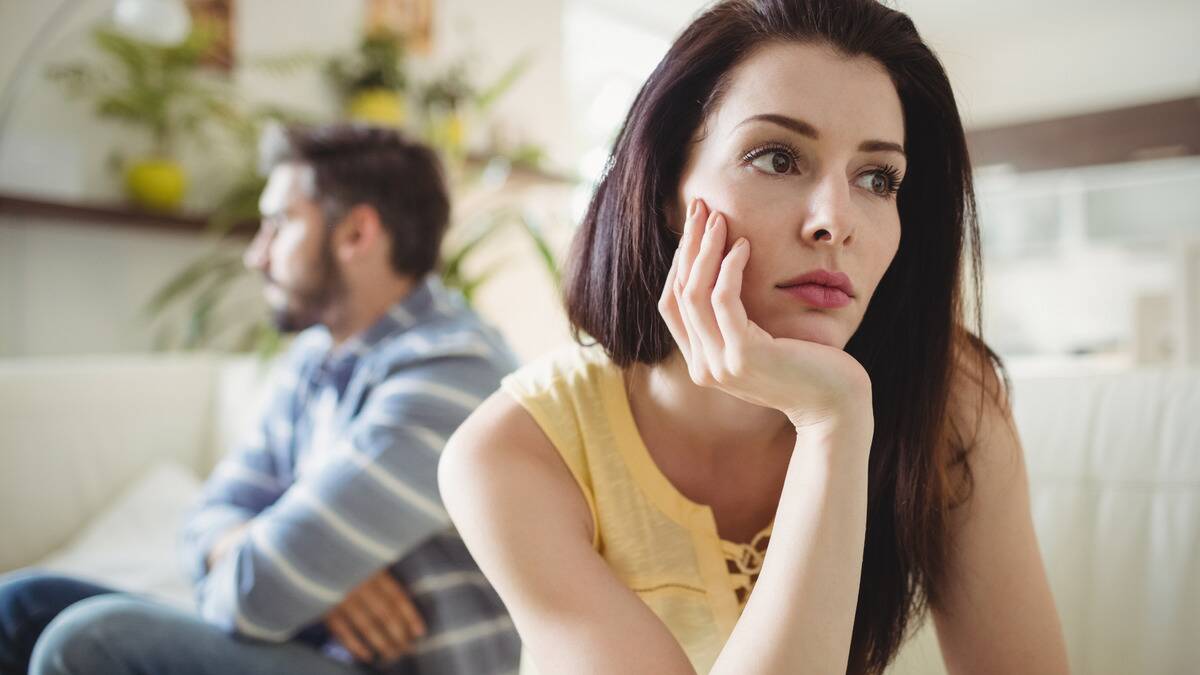 A woman with her chin in her hand, looking away sadly, as a man behind her sits with his arms crossed.