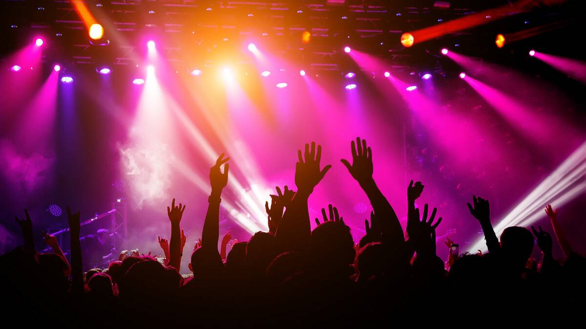 A crowd at a music venue silhouetted by the bright pink, purple, and orange stage lights, hands seen raised in the air.