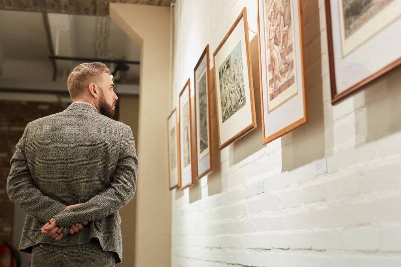 A man walks with his hands behind his back, looking at art hung on the wall.