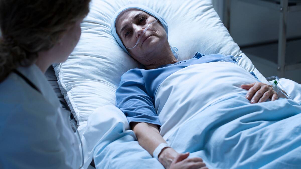 An ill woman laying in a hospital bed, looking up at the doctor speaking to her.