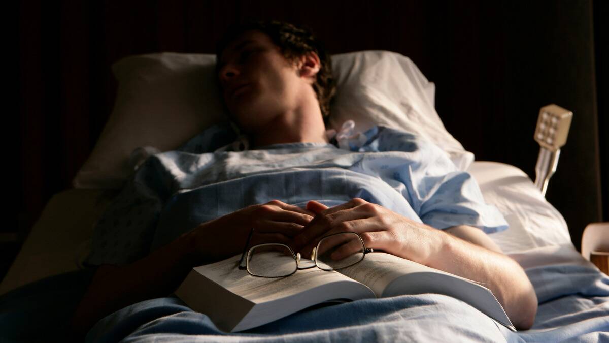 A man laying in a hospital bed at night, having fallen asleep, open book and glasses resting on his lap.