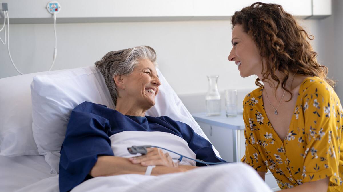 A woman in a hospital bed smiling as she speaks to someone, presumably her daughter, sitting next to her.