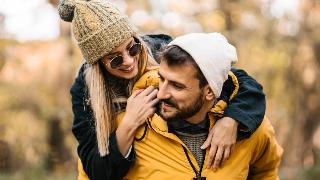 A couple walking through an autumn forest, the man giving the woman a piggyback ride, both smiling.