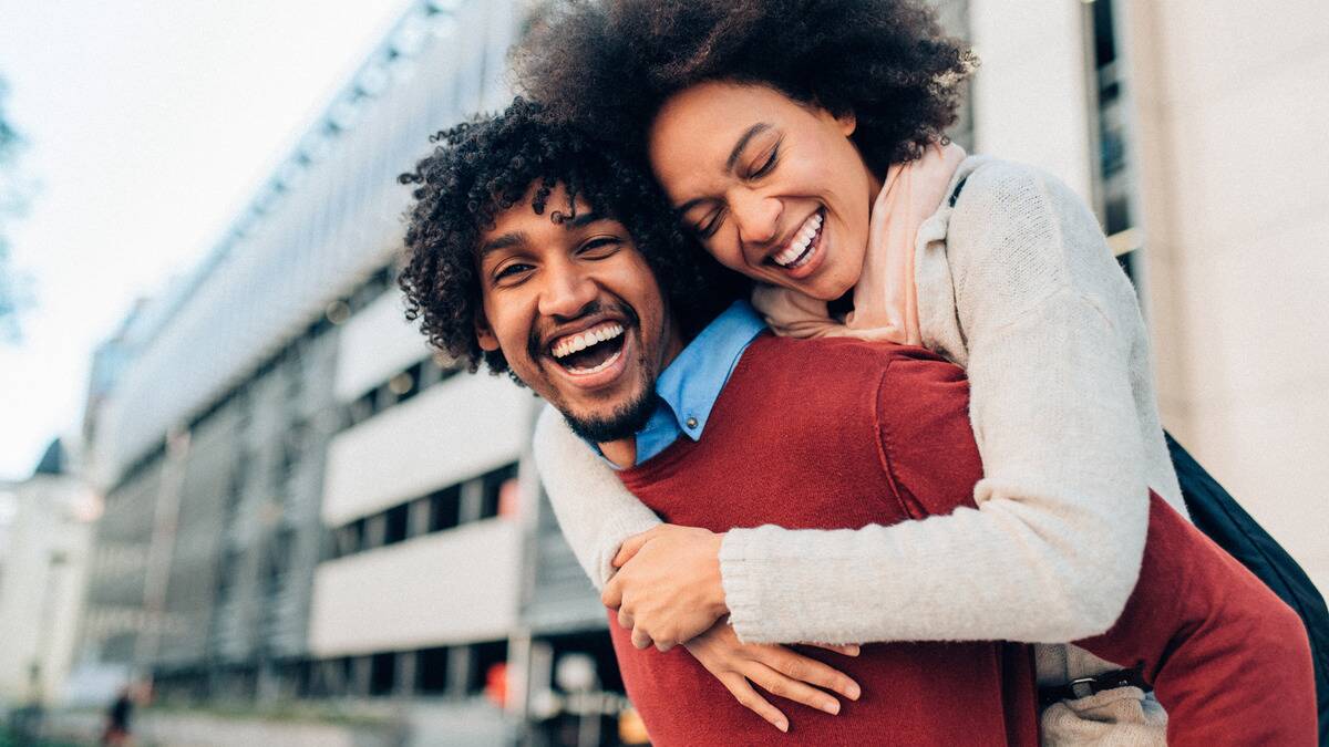 A couple walking through a city street, both laughing as the man gives the woman a piggyback ride.