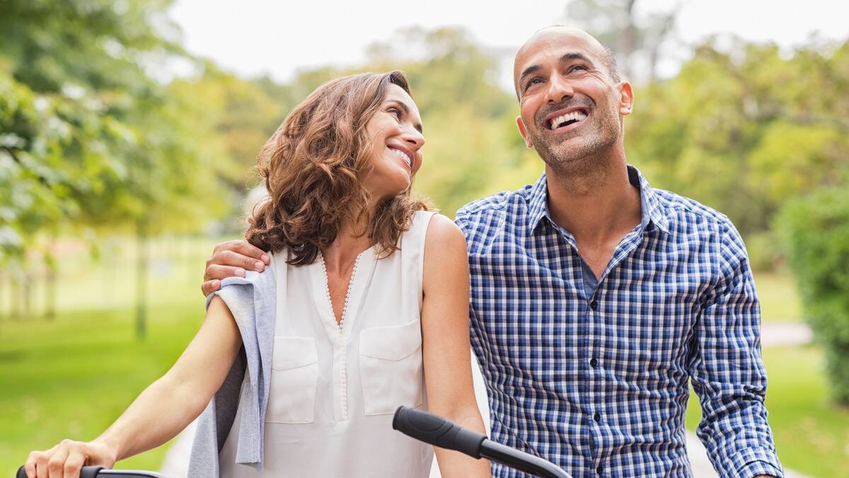 A couple walking through a park, both leading their bikes, the man with his free arm around the woman's shoulder, both smiling.