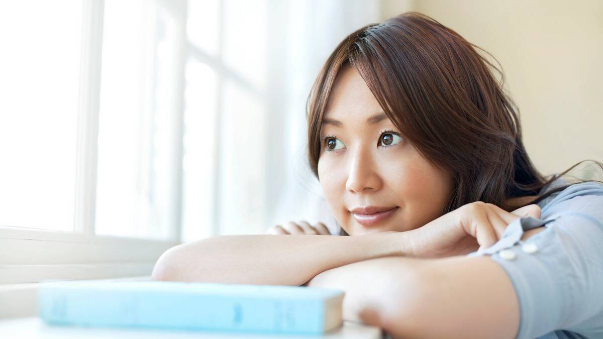 A woman with her arms folded on a table, resting her chin on them, looking out a window.