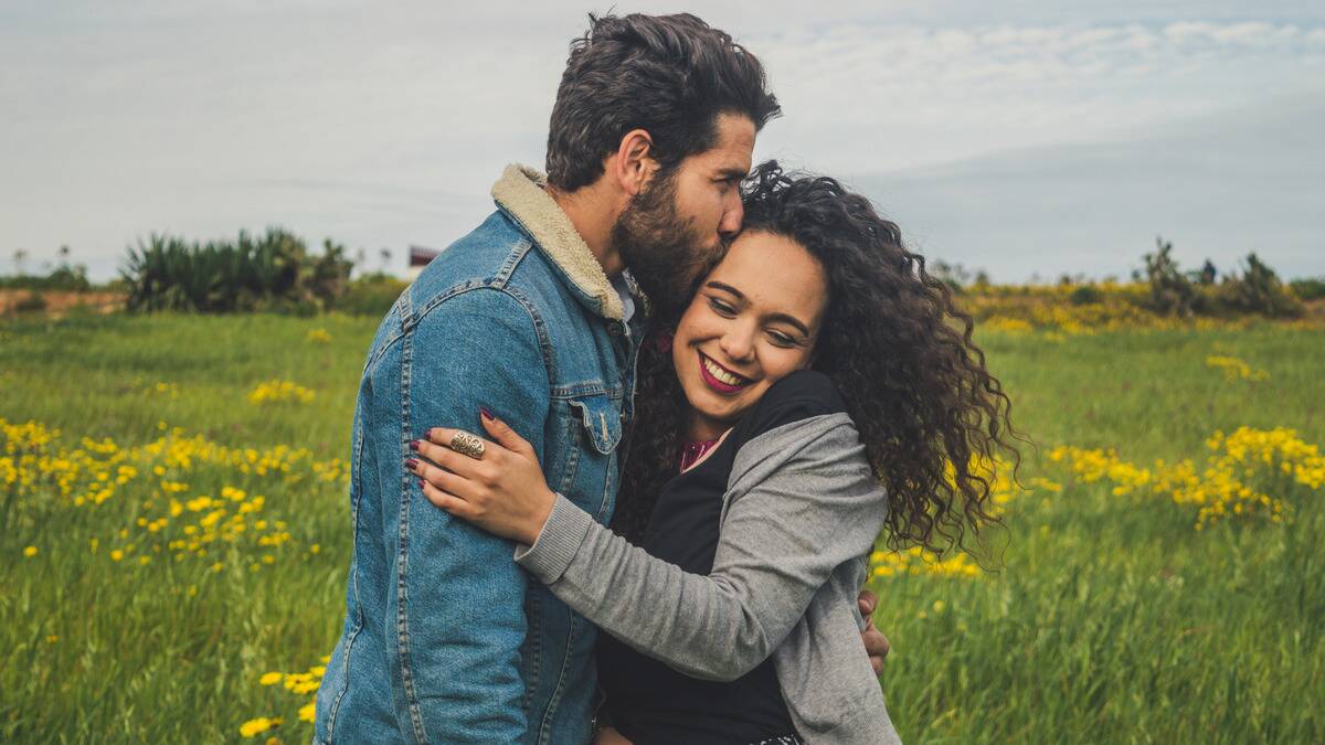 A couple standing outside, arms around one another, the man kissing the woman's temple as she smiles.