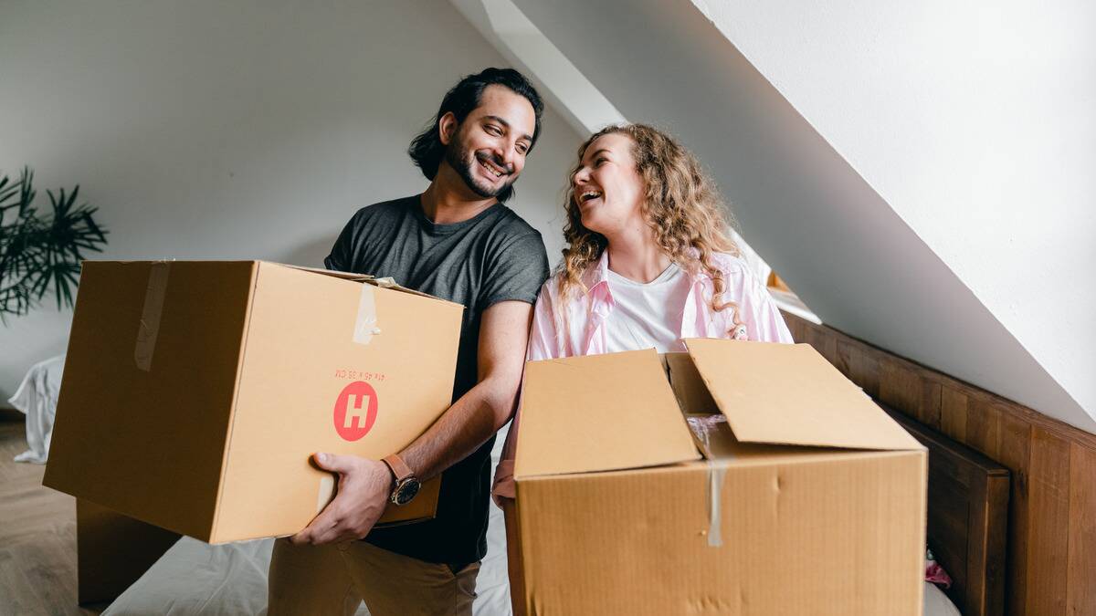 A couple each holding up a moving box, smiling at one another.