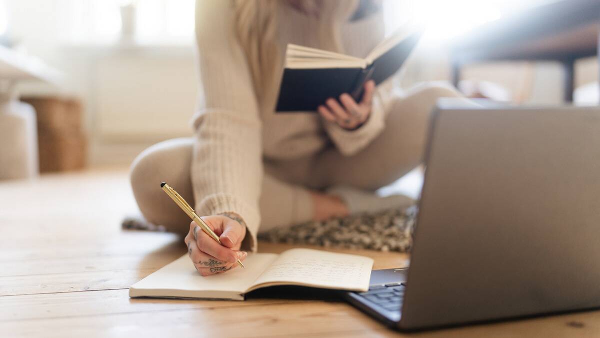 A woman sitting on the floor, holding a small book, laptop open in front of her, writing in a separate notebook.