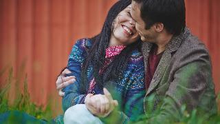 A couple sitting side by side, arms around one another, faces close, smiling.