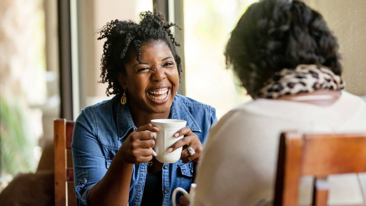 A woman smiling as she chats to a friend across a cafe table, holding up a mug.