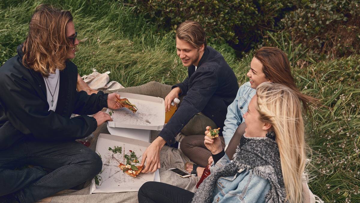 Friends sat around a pizza box on a picnic blanket, smiling as they chat and eat.