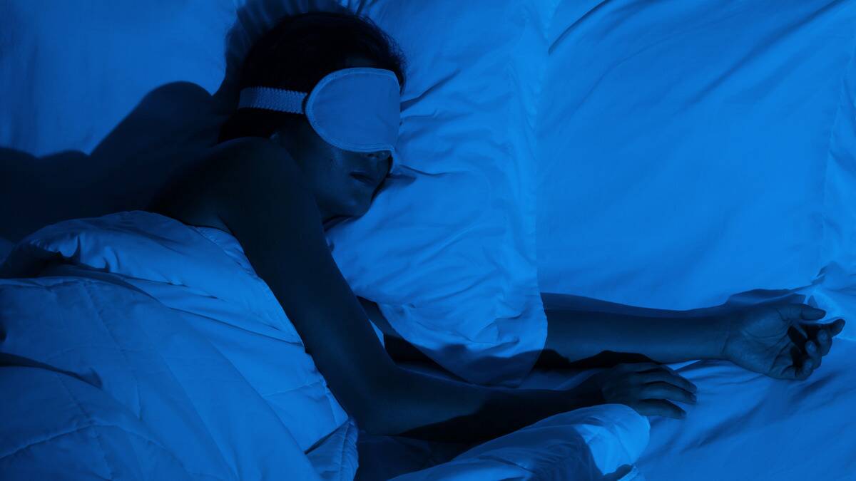 A blue-lit shot of a woman sleeping in bed on her side, arms sprawled out, eyemask on.