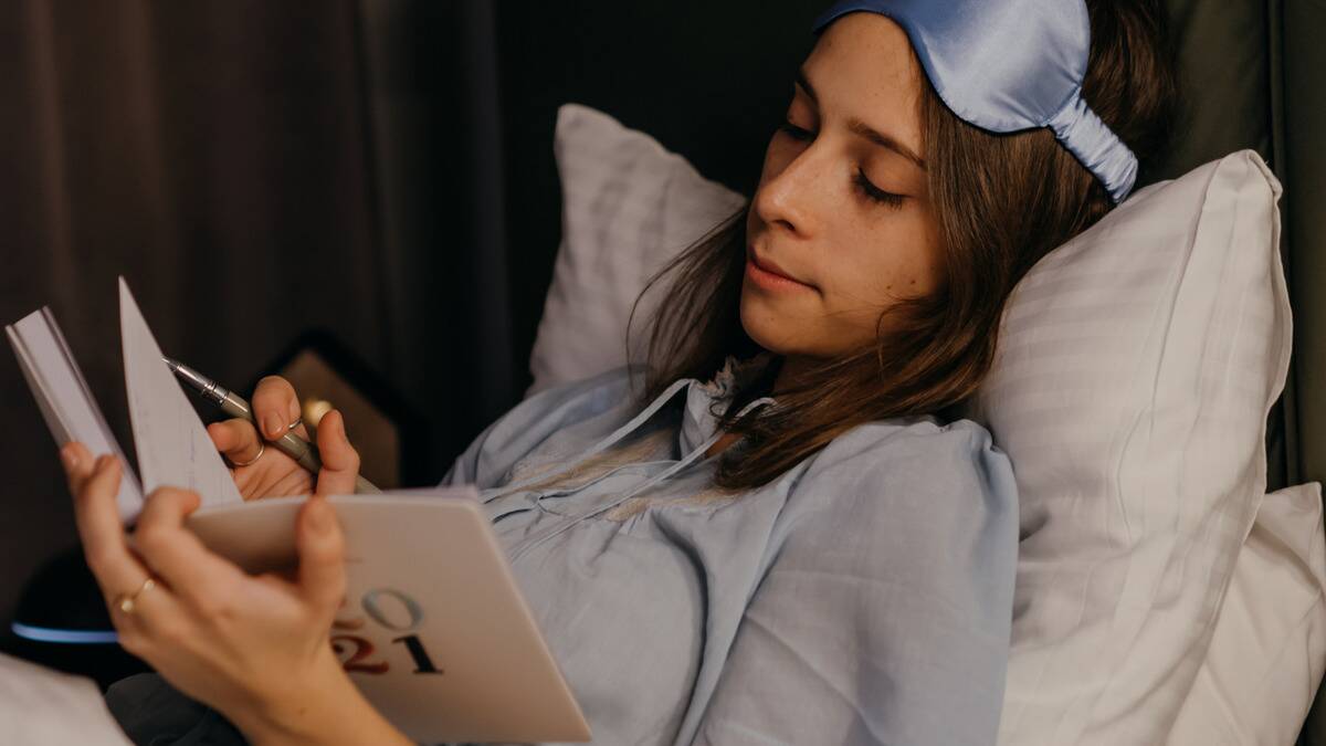 A woman sitting up in bed, eyemask pulled onto her forehead, writing in a journal.