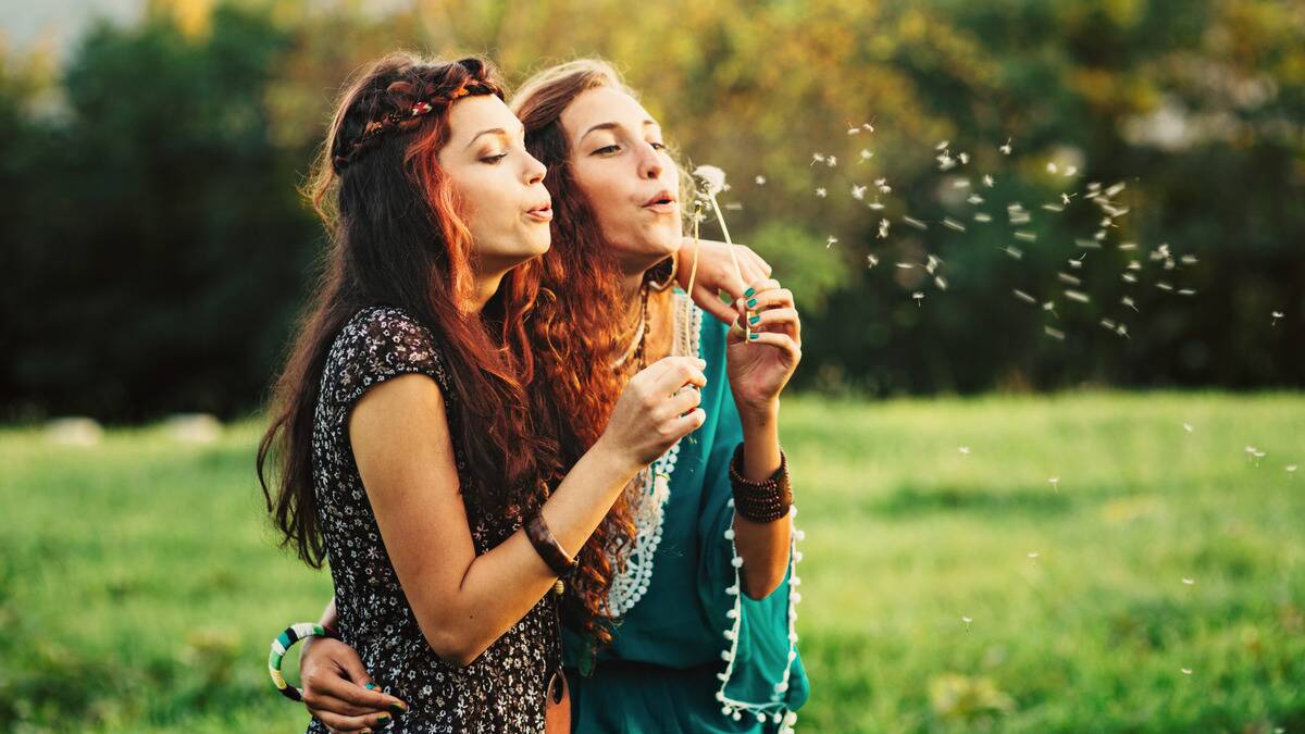 Two friends standing next to each other with their arms around each other, blowing the seeds of a dandelion.