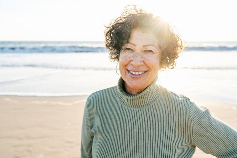 A kind woman smiling on the beach, one hand on her hip.