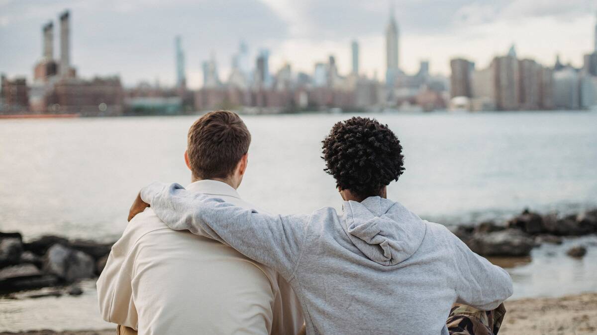 Two friends sat by the waterfront looking at a cityscape, one with his arm around the other.