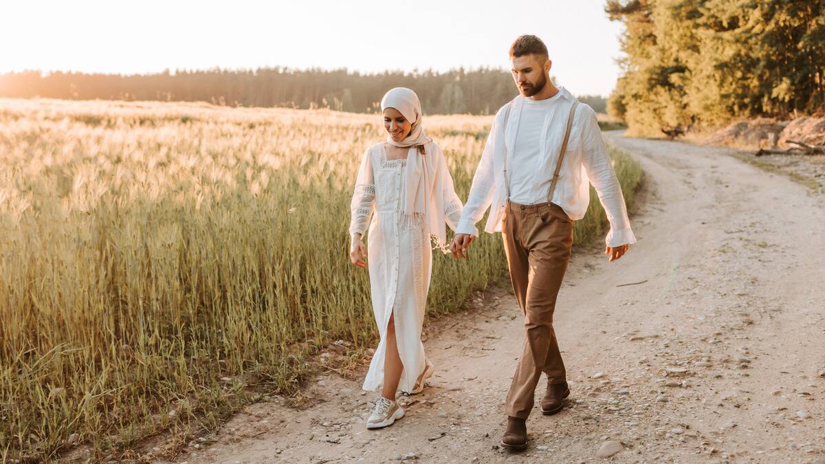 A couple holding hands as they walk along a dirt road next to a field.