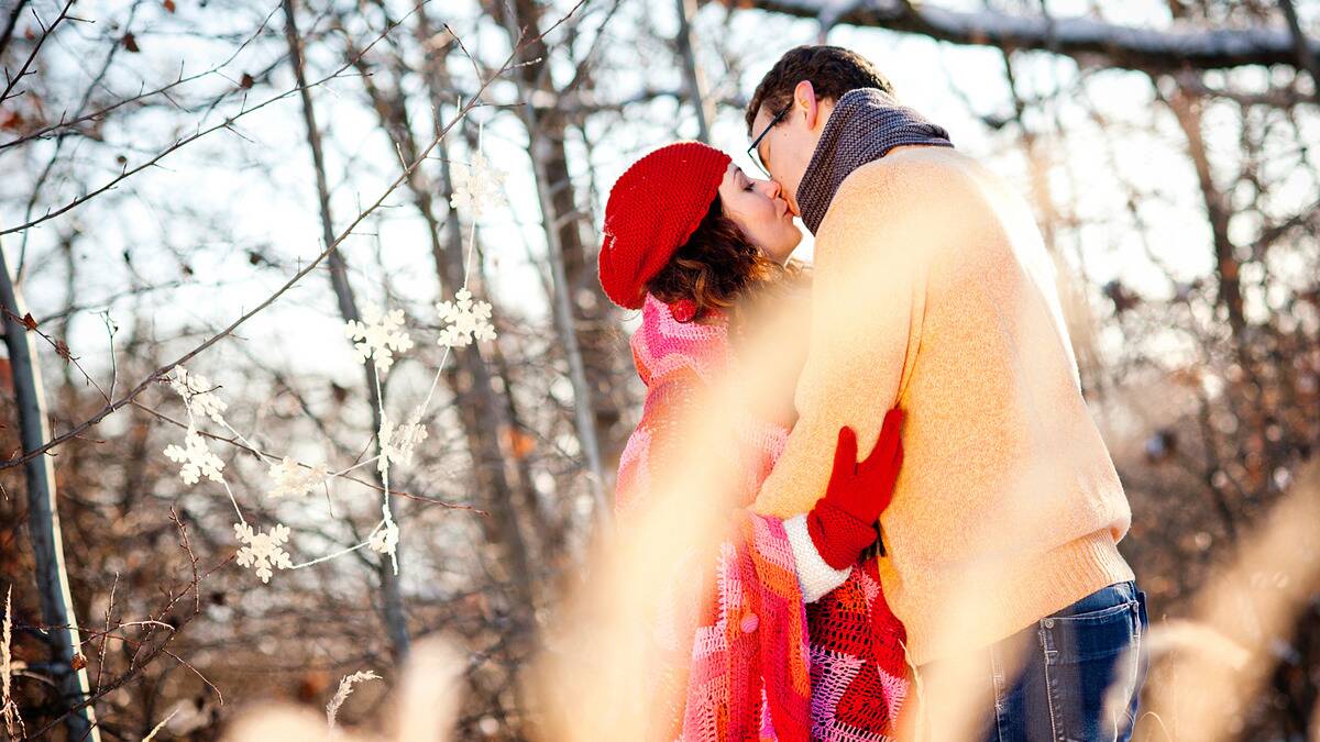 A couple standing outside in the winter, kissing as they stand among the trees.