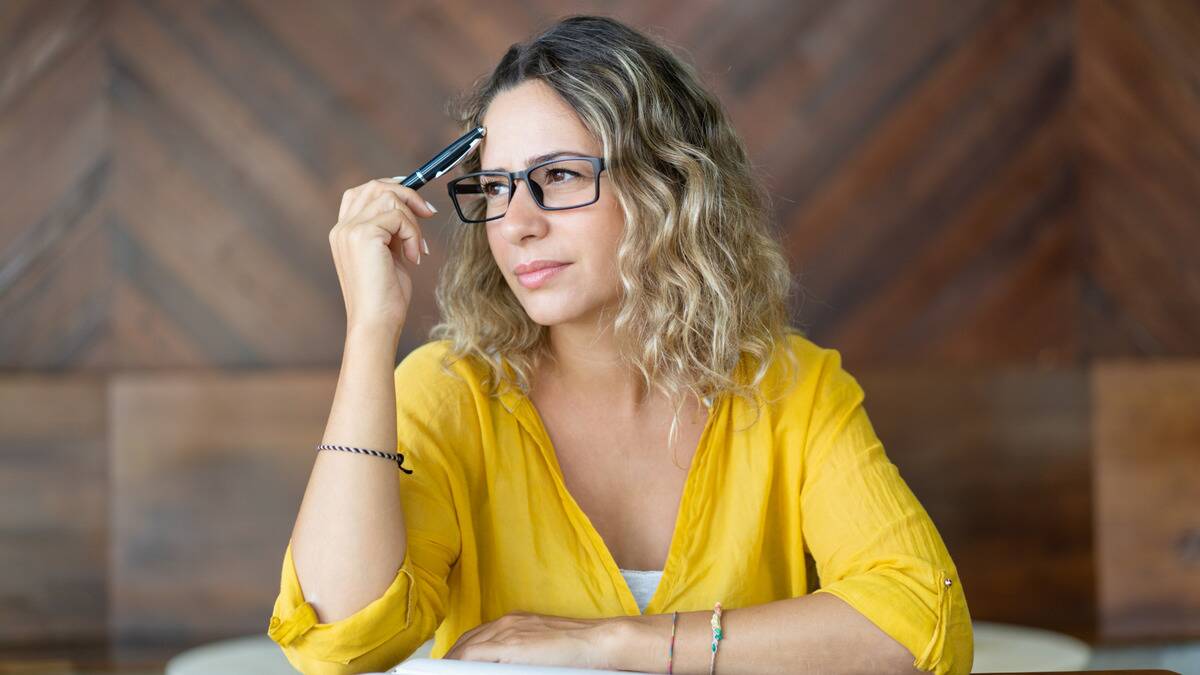 A woman sitting at a table, tapping a pen to her forehead, looking pensive.