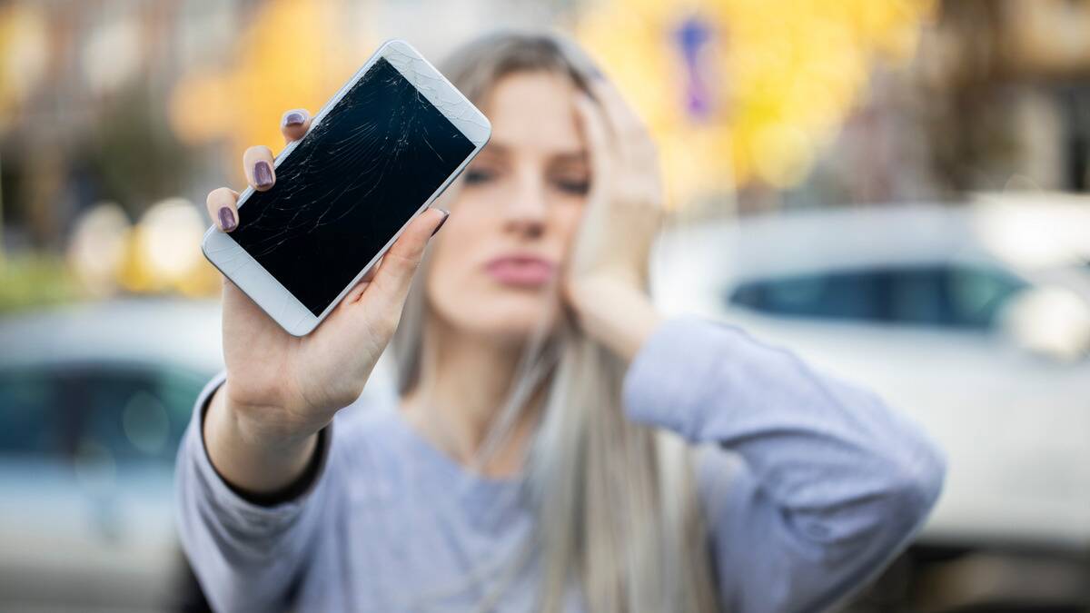 A woman holding up her phone, showing off the cracks on the screen, her face visible in the background as she holds her head with her other hand.