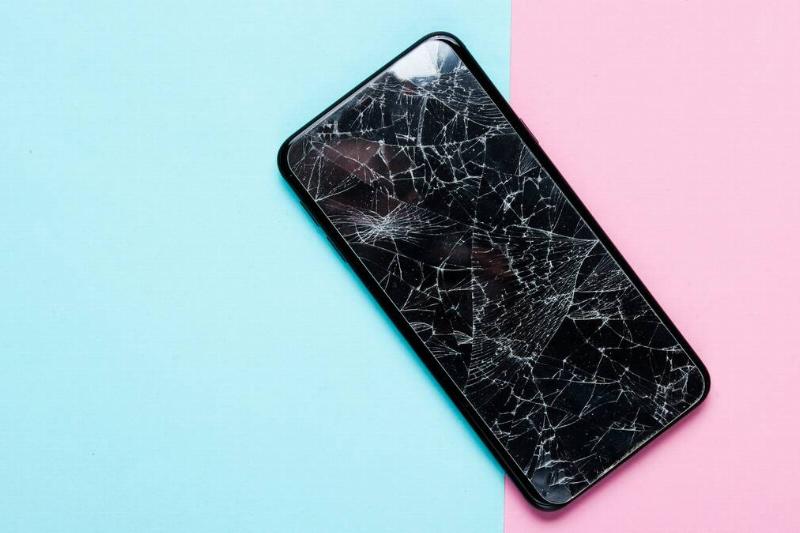 A phone with an extremely cracked screen laying on a pink and blue background.
