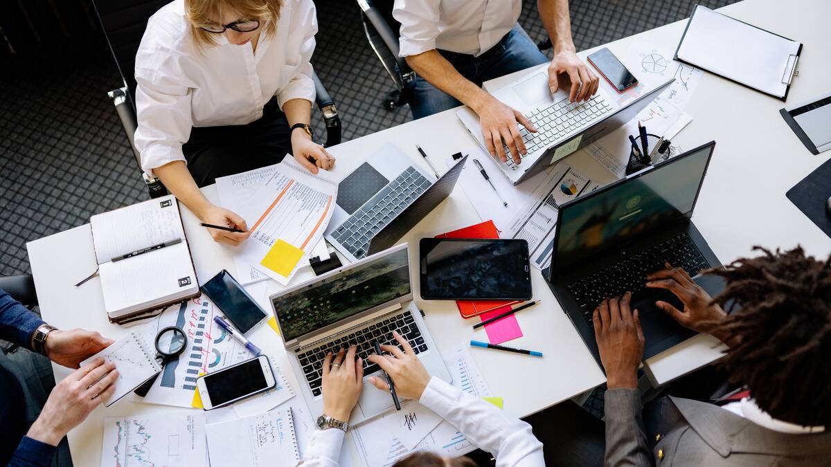 An aerial shot of a desk with five people sat around it working, laptops and documents strewn about.