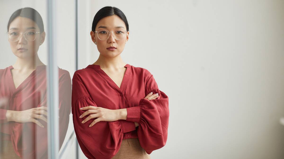 A woman leaning on a glass wall with her arms crossed, looking at the camera sternly.