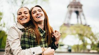 Two friends hugging and smiling together, the Eiffel Tower seen blurred in the background.