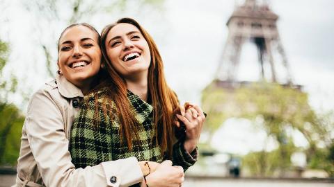 Two friends hugging and smiling together, the Eiffel Tower seen blurred in the background.