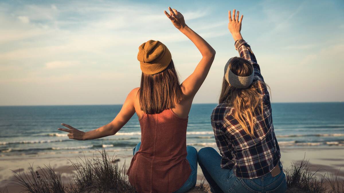 Two friends sitting side by side near a beach, both with their hands raised to the sky.