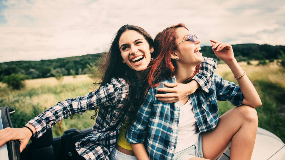 Two friends laughing as they sit together by a field, one with her arm around the other.