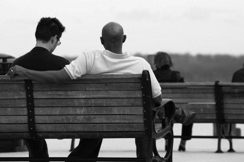 A greyscale image of two friends sitting on a park bench, one with his arm over the back of the bench.