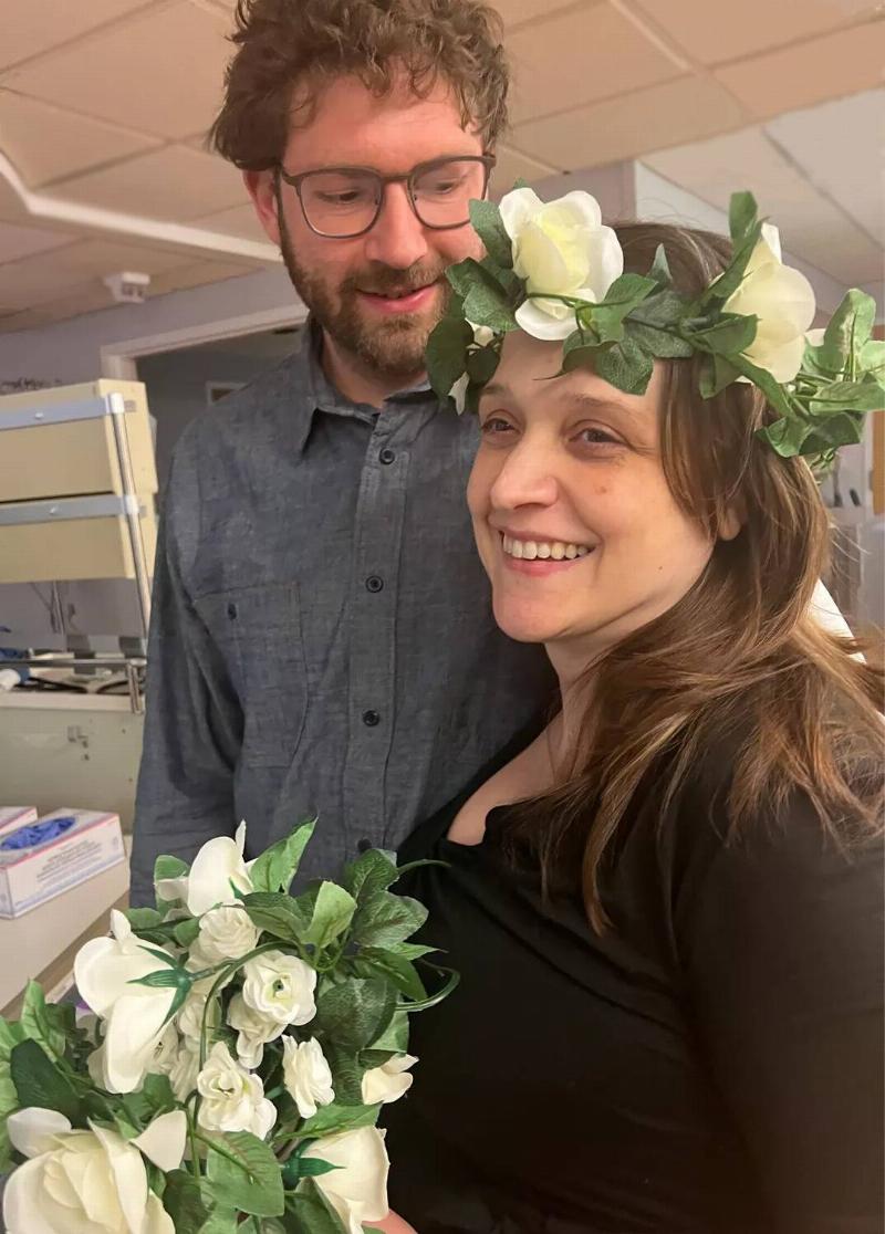 Nora and Michael standing in the hospital before their marriage, Nora in a flower crown while holding a bouquet made by the nurses.