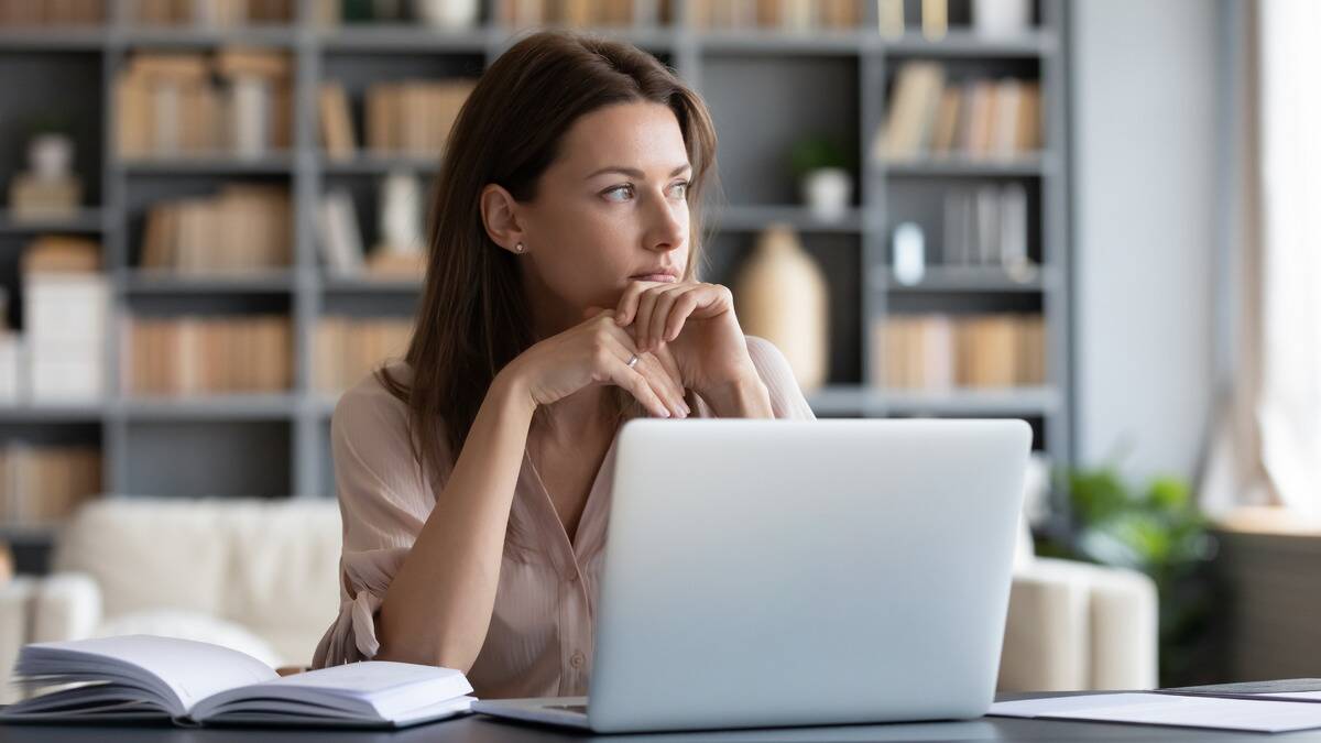 A woman sitting at her desk, laptop and book open in front of her, but she's looking off to the side, distracted.