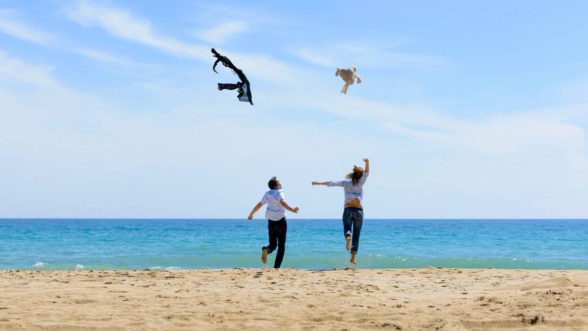 Two people running toward the water on a beach, each throwing their jackets up into the air.