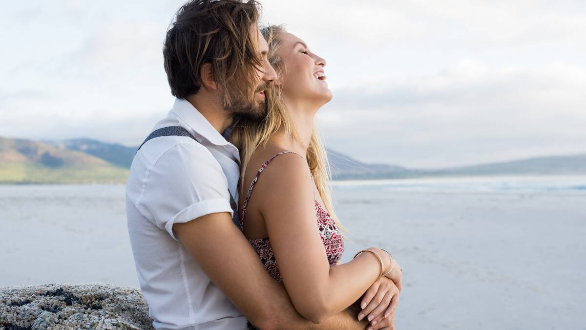 A couple standing by the water, the woman in front of the man, smiling as she leans back into his chest and he puts his arms around her.