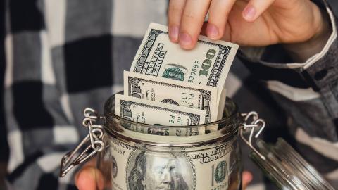 A close shot of someone pulling a $100 bill from a jar full of $100 bills.