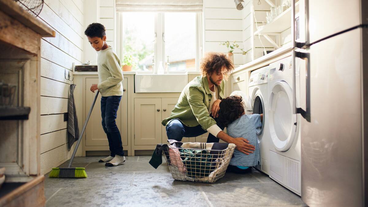 A father kneeling on the floor helping teach his young son how to do laundry. Behind him, his other son is helping sweep the floor.