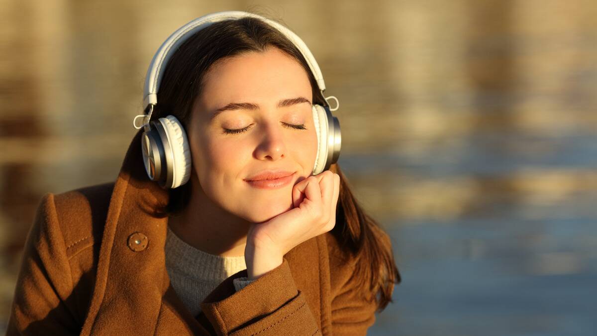 A woman sitting by the water, wearing headphones, resting her chin in her hand, eyes closed with a peaceful smile.