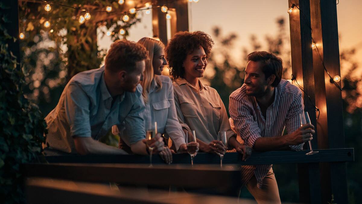 A group of four friends leaning against a deck railing, all smiling as they chat, glasses of champagne in hand.