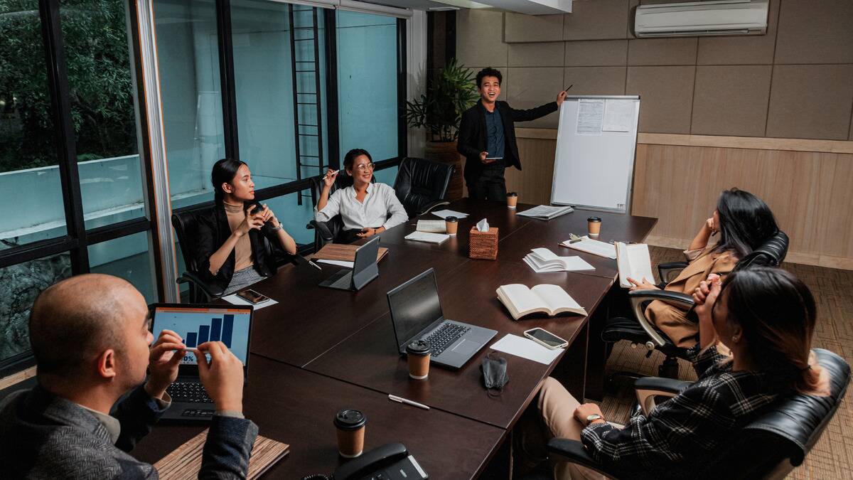 A workplace meeting around a boardroom table, one man standing next to a white board as he speaks.